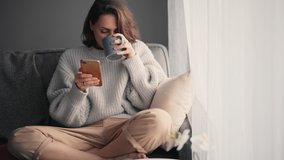 Relaxed young woman using a smartphone and drinking coffee from a mug while sitting on the sofa. Millennial girl spending time at home with cell gadget technology. - Powered by Shutterstock - Get 15% off with code: PIKWIZARD15