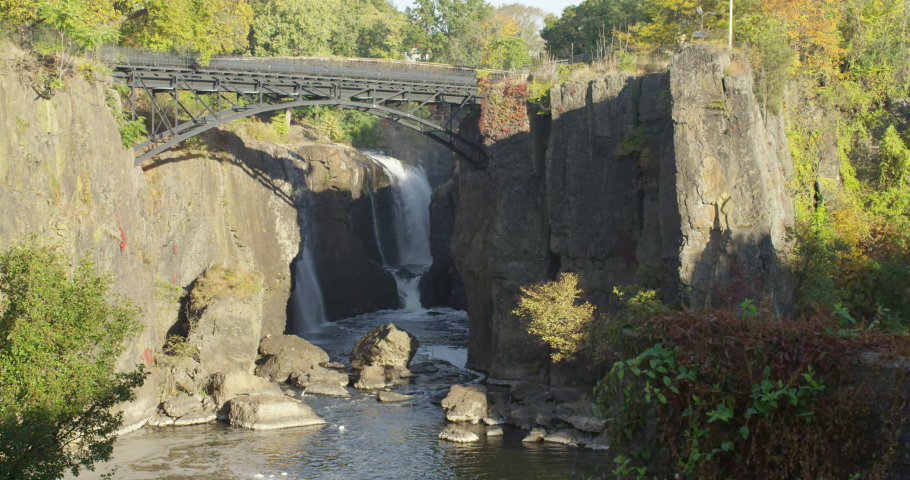 A huge waterfall in a city park with a footbridge over the cliffs during sunny autumn morning, slow motion