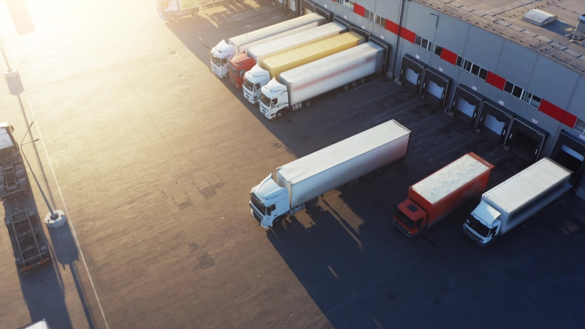 Trucks with semi-trailers stand on the parking lot of the logistics park with loading hub and wait for load and unload goods at warehouse ramps at sunset. Aerial view Royalty-Free Stock Footage #1063979203
