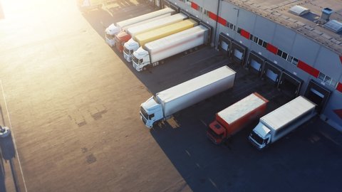 Trucks with semi-trailers stand on the parking lot of the logistics park with loading hub and wait for load and unload goods at warehouse ramps at sunset. Aerial view