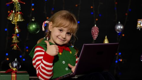Child looking for presents, gift for friends and family, doing shopping online using laptop isolated on coronavirus quarantine lockdown. Kid girl in Christmas elf Santa Claus helper costume. 