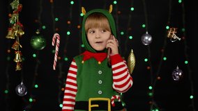 Joyful kid child girl in Christmas elf Santa helper costume making call on mobile phone to Santa Claus or sends messages of congratulations, online gifts. Black background. 