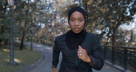 Front view of young pretty muslim woman running in park. Energetic female person in hijab and sports suit jogging while looking to camera. Concept of healthy lifestyle, sport. Outdoors.