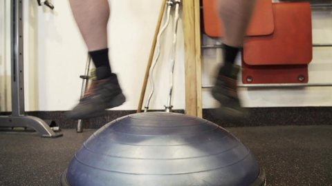 Strong Muscular Man in T-shirt and Shorts Exercises Jumping on a Bosu Ball in his home gym, close up