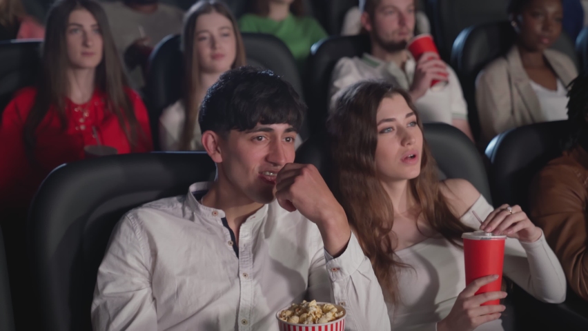 Motion of young gorgeous woman watching film in cinema with boyfriend eating popcorn, audience on background. Attractive caucasian female enjoying film holding fizzy drink, multiracial audience.