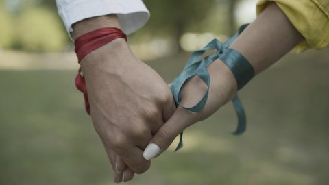 Close-up of male and female Caucasian hands with ribbons in sunlight. Unrecognizable hippie man and woman holding hands outdoors. Pacifism and 1960s lifestyle.