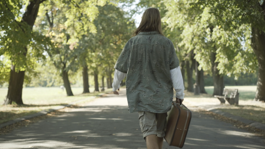 Back view of carefree hippie man walking along the road on sunny summer day and looking around. Relaxed Caucasian young man strolling outdoors in 1960s. Pacifism and lifestyle concept.