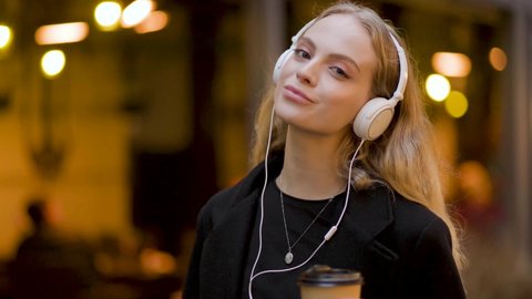Slow motion of happy young woman in headphones walking outdoors in city street having fun alone. Joyful attractive blonde carefree woman listening to music and drink coffee