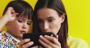 Closeup adorable little son and mother holding cellphone watching funny video clips or playing in game on phone. Laughing family resting together isolated on yellow background.
