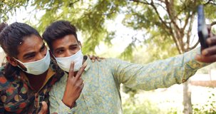 Slow-motion static shot close-up portrait of two young male adult brothers, wearing face mask in nature park, take selfie photo video using a smart phone mobile as they smile and make V hand sign