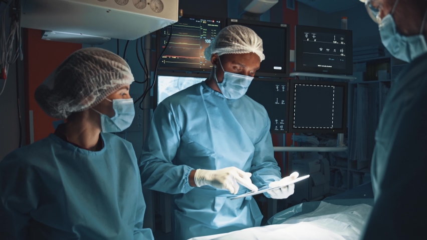 Healthcare and modern medicine. Serious multirace doctor looking together with his team at X-ray and CT results showing at digital tablet while preparing at the serious operation at operation room | Shutterstock HD Video #1063997434