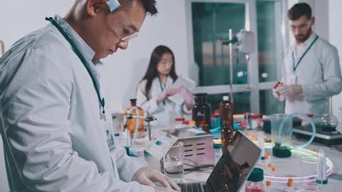 Asian scientist using laptop computer while collaborating in laboratory. Multi-ethnic professional lab workers developing medications in research facility center.