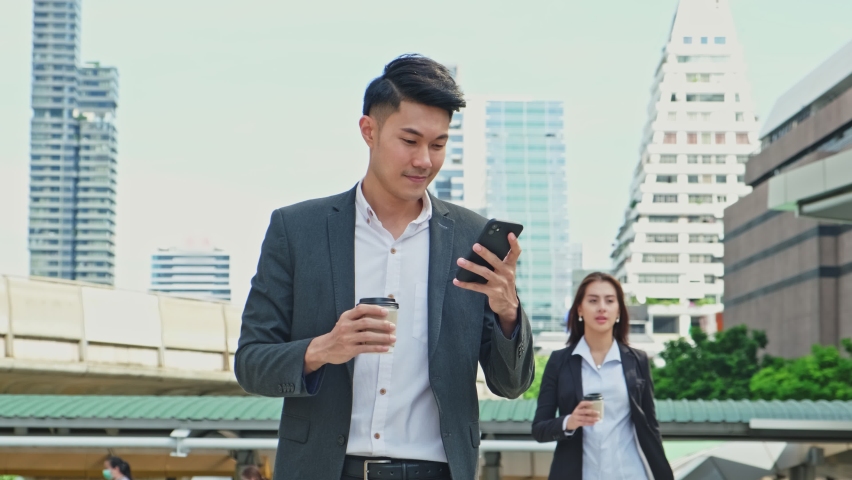 Asian young smart businessman using smartphone in city. The man holding phone walking outdoor to working office with smile face. Telecommunication and mobile network for business concept. Royalty-Free Stock Footage #1063999291