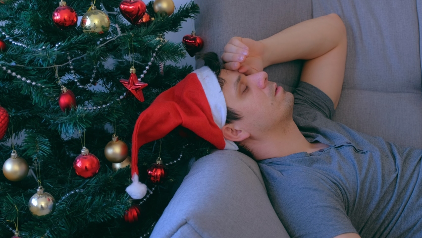 Portrait of man sleeping lying on couch at home near Christmas tree at eve in red cap, side view. Morning hangover after party. Celebrating holiday. He is napping waiting 12 night. He is snores.