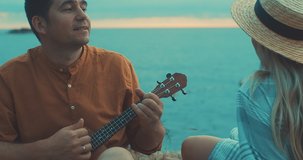Fun attractive couple of traveler sitting on sea beach with ukulele under sunset sky in evening time. Man playing, woman listening and sing music, relaxing enjoying holidays, travel day. 4K video