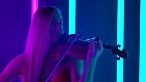 Detailed shot of a young woman musician masterly playing the violin. The blonde touches the strings with a bow close up against the background of bright neon lights. Slow motion.