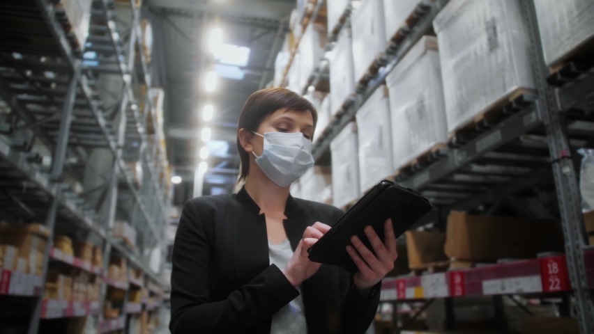 Warehouse supervisor checking quality and availability of goods by gadget. Staff of warehouse wearing safety mask to protect coronavirus. E-commerce. Online delivery. Reopening after pandemic. Royalty-Free Stock Footage #1064011147