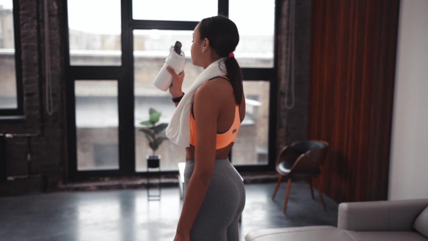 Athletic African American beautiful woman drinking protein shake or water and wiping sweat off her face with a towel, workout in a cozy home environment. Royalty-Free Stock Footage #1064013919