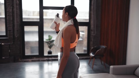 Athletic African American beautiful woman drinking protein shake or water and wiping sweat off her face with a towel, workout in a cozy home environment.