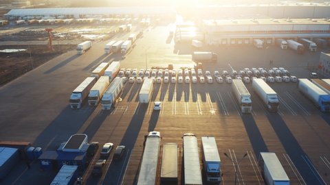 Semi-trailer trucks stand on the parking lot of the logistics park with loading hub and wait for load and unload goods at warehouse ramps at sunset. Aerial view