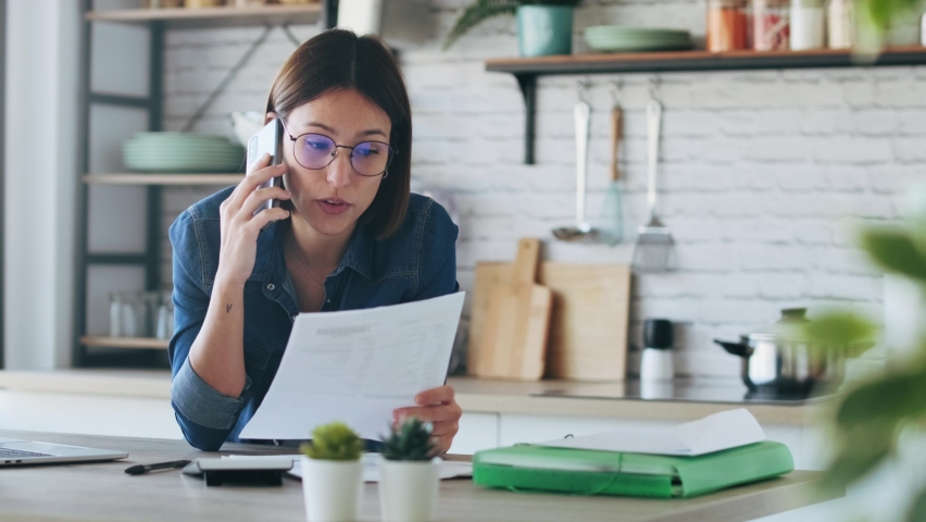 Video of beautiful young business woman talking with her mobile phone while consulting some invoices and documents in the kitchen at home. Royalty-Free Stock Footage #1064017924