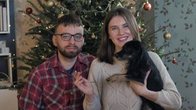 View from camera: happy caucasian couple with little cute black dog record video of New Year's greetings for friends and family standing at home near Christmas tree. Christmas congratulation via web