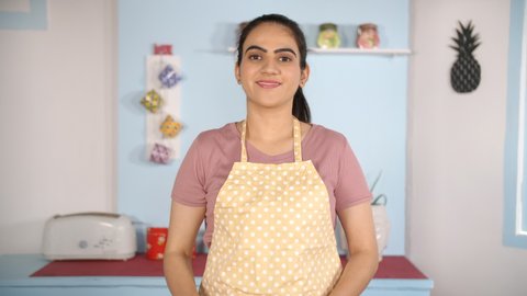 Smiling young woman wearing a printed apron while standing with her folded arms. Medium shot of a confident modern home chef holding a wooden spatula while standing in the kitchen
