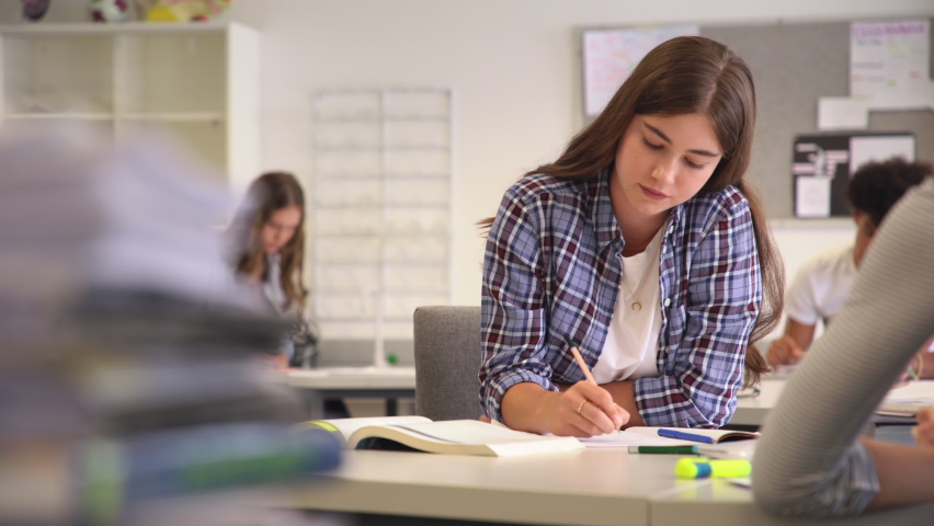 Beautiful university student smiling while studying for exams in classroom. Pretty woman sitting in classroom full of students during class. Portrait of happy young woman writing notes. | Shutterstock HD Video #1064040595