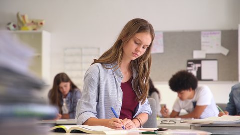 Portrait of smiling high school student with classmates in background writing notes in the classroom. Happy casual girl sitting at desk in class while looking at camera.