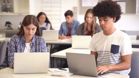 College students sitting together at desk and using laptop during computer lesson. Young black man and casual girl working on laptop in high school library. Happy smiling multiethnic friends working.