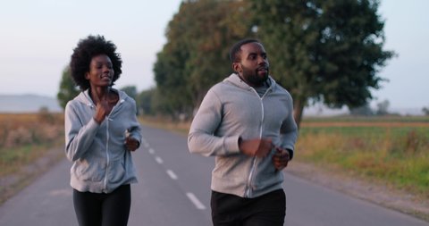 African American young sporty man and woman joggers running on road early in the morning in countryside. Couple of runners. Sportswoman and sportsman jogging in nature.