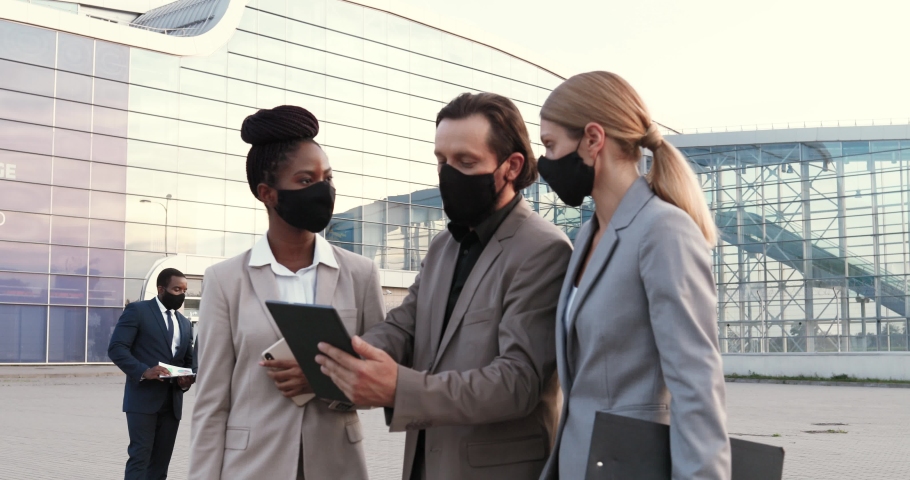 Mixed-races young male and females business partners in masks talking and using tablet device outdoor at street. Businessman and businesswomen having conversation in city. Pandemic coronavirus.