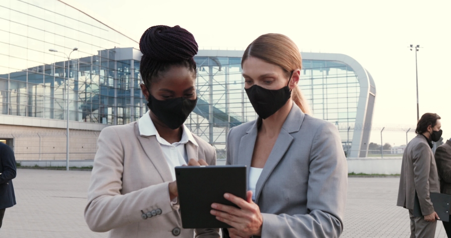 Mixed-races young females business partners in masks talking and using tablet device outdoors at street. Multi ethnic businesswomen having conversation in city. Pandemic coronavirus. Women colleagues.
