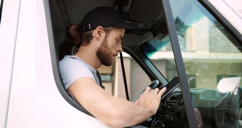 Side view of young Caucasian male courier worker sitting on front passenger seat in van typing and browsing online on tablet alone. Close up of delivery man using device. Shipment service concept