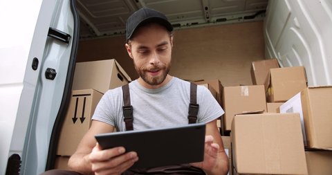 Close up portrait of Caucasian happy young male courier sitting in van with many boxes tapping and browsing on tablet device in good mood at work. Delivery man delivering shipment. Shipping concept