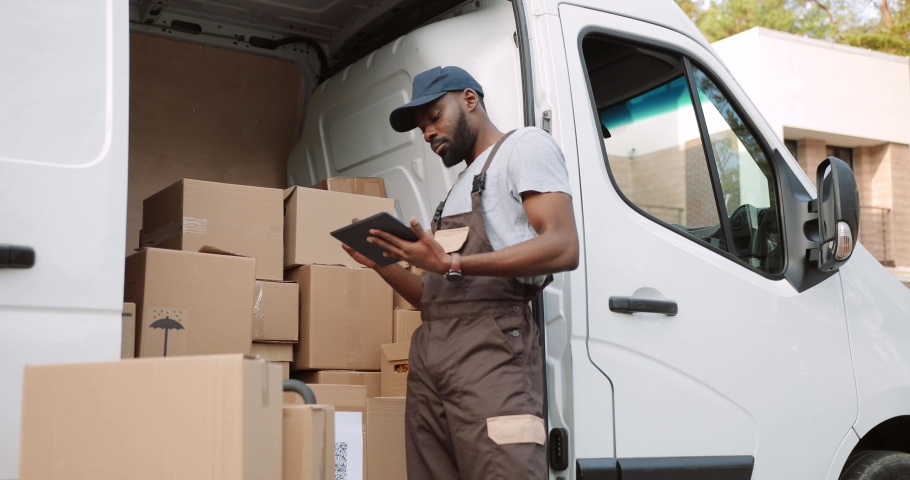Young handsome African American delivery man typing and tapping on tablet device standing near van on street with many ordered boxes. Male postman courier with shopping package. Shipment concept Royalty-Free Stock Footage #1064056555