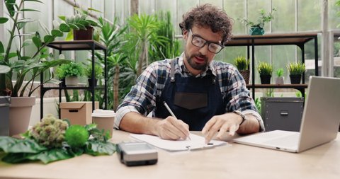 Portrait of handsome man in glasses and apron looking at laptop and writing down in planner while sitting in cozy floral shop. Caucasian young male florist flower store owner working. Business concept