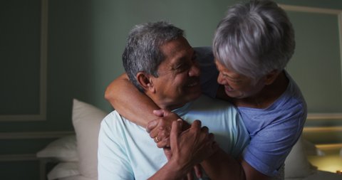 Senior mixed race couple embracing and laughing in bedroom. self isolation retirement lifestyle at home during coronavirus covid 19 pandemic.