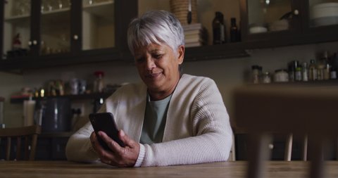 Smiling senior mixed race woman using smartphone sitting in kitchen. self isolation retirement lifestyle at home during coronavirus covid 19 pandemic.