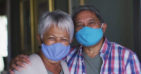 Senior mixed race couple wearing face masks standing embracing in hallway. hygiene health self isolation retirement lifestyle at home during coronavirus covid 19 pandemic.
