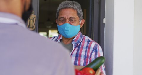 Senior mixed race man wearing face mask taking food delivery. hygiene health self isolation retirement lifestyle at home during coronavirus covid 19 pandemic.