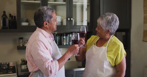 Happy senior mixed race couple wearing aprons toasting with wine in kitchen. self isolation retirement lifestyle at home during coronavirus covid 19 pandemic.