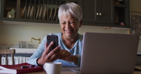 Happy senior mixed race woman using smartphone and laptop in kitchen. self isolation retirement lifestyle at home during coronavirus covid 19 pandemic.