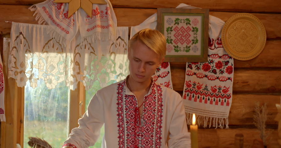Portrait, a Russian Slavic man in a folk costume sits at a table in a hut, writes with a pen in a book. Decoration of the Russian hut, curtains and a tablecloth with embroidery. 4k, ProRes