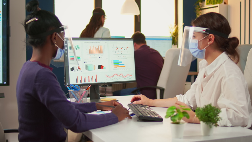 Office manager explaining to black colleague financial startegy pointing graphs on desktop wearing face masks and visors. Multiethnic team working in company with new normal respecting social distance - Powered by Shutterstock - Get 15% off with code: PIKWIZARD15