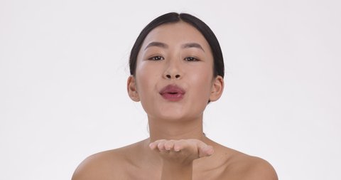 Young asian woman sending blow kiss to camera, posing over white studio background, slow motion, beauty portrait