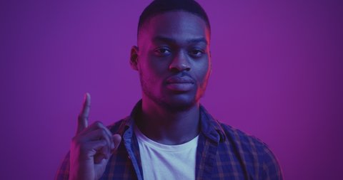 BLM. Young serious black guy turning face to camera and pointing finger up, looking at camera in neon lights background, close up