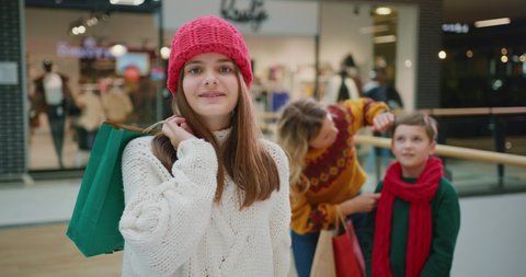 Young smiling caucasian girl holding bag with Christmas gift posing at camera in shopping mall. Her family mother and brother standing in background. Holiday shopping.