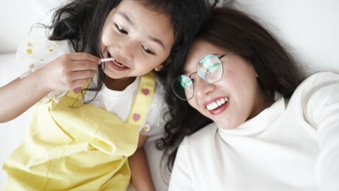 Young Asian mother with a little child holding phone while taking video calls with dad is working in another country on the bed in bedroom. Happy child smiles and laughs with mother playing together.