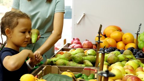 Woman with little daughter is choosing a fresh fruits at grocery store together. Mother with child girl is taking ripe pears from shelf and puts it in shopping basket